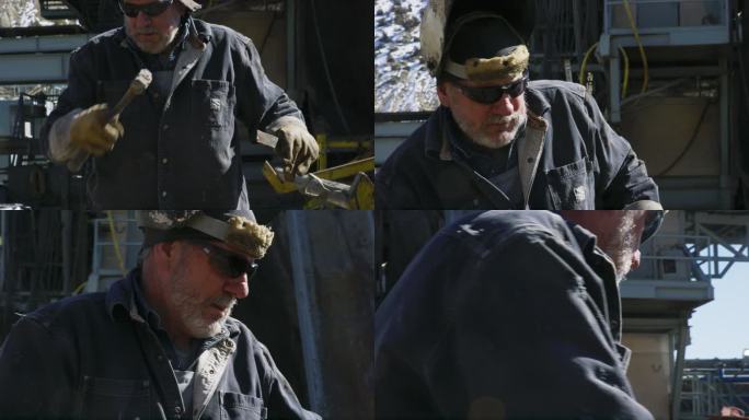阳光明媚的一天，一名60多岁的油田男工人戴着焊接面罩，在石油天然气钻井平台现场的井架旁拿着锤子和锉刀