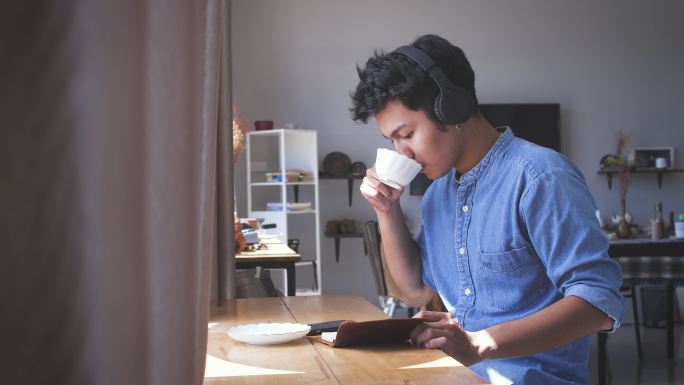 年轻的亚洲帅哥在家里一边看书一边用耳机听音乐，一边喝咖啡，人们放松的生活方式和科技理念