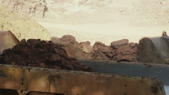 阳光明媚的施工现场，挖掘机铲斗将泥土和岩石倒入自卸车后部的特写镜头（铰接式自卸车）