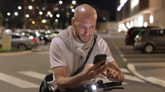 年轻人晚上在城里用手机开着电动滑板车