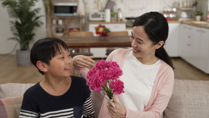 亚洲男孩在家里给妈妈送母亲节花的后视图。微笑的女人轻轻地抚摸儿子的脸，闻着花束，一边说谢谢
