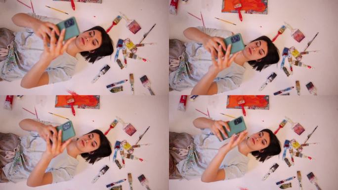 无忧无虑的女艺术家躺在画布上使用手机