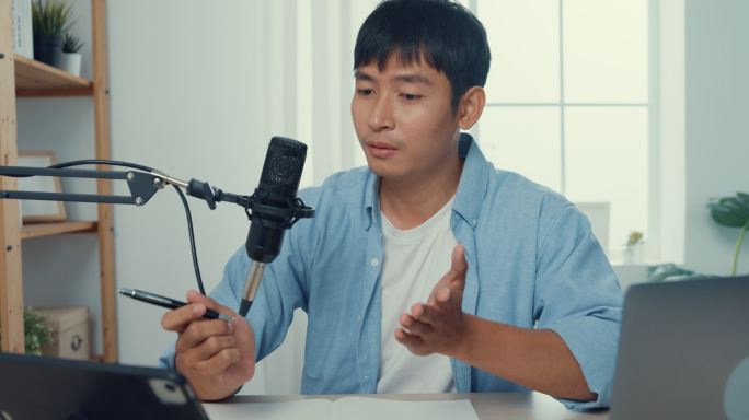 一位年轻的亚洲男子坐在家里客厅的工作台上，用显微镜在她的笔记本电脑上录制播客。