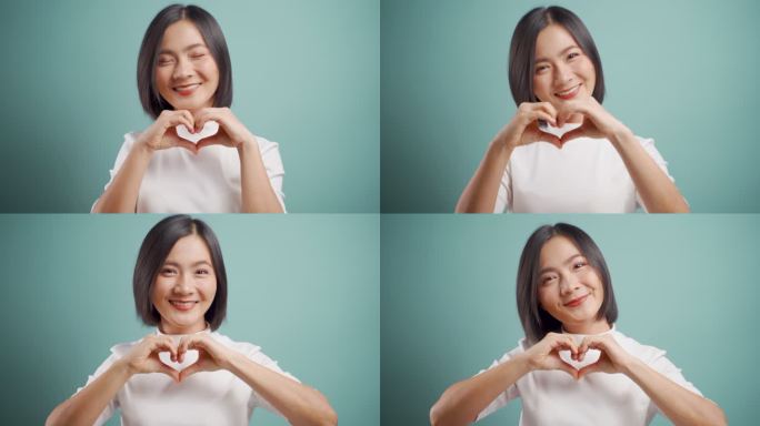 亚洲女性在恋爱中开心地微笑，用手塑造心形，看着孤立地站在蓝色背景上的相机。4K视频。情感上的幻想。