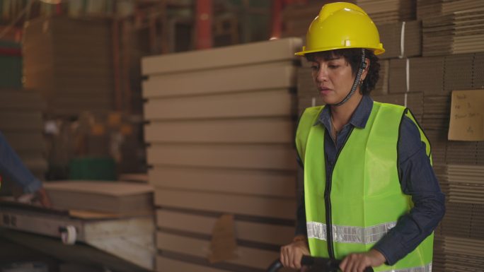 在造纸厂，一名穿着安全背心和头盔的拉丁妇女纸包装制造工人使用手动液压托盘车移动纸板堆
