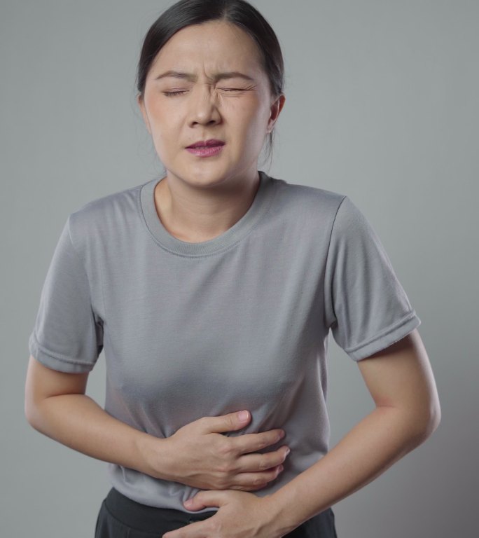 一位亚洲女性因胃痛而病倒，她站在灰色的背景上，独自一人。垂直视频4k分辨率。