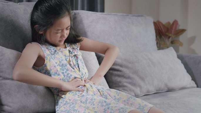 亚洲女孩因食物中毒而胃痛。她坐在家客厅的沙发上，用手捂住胃痛。一个女孩在吃快餐后身体受伤。
