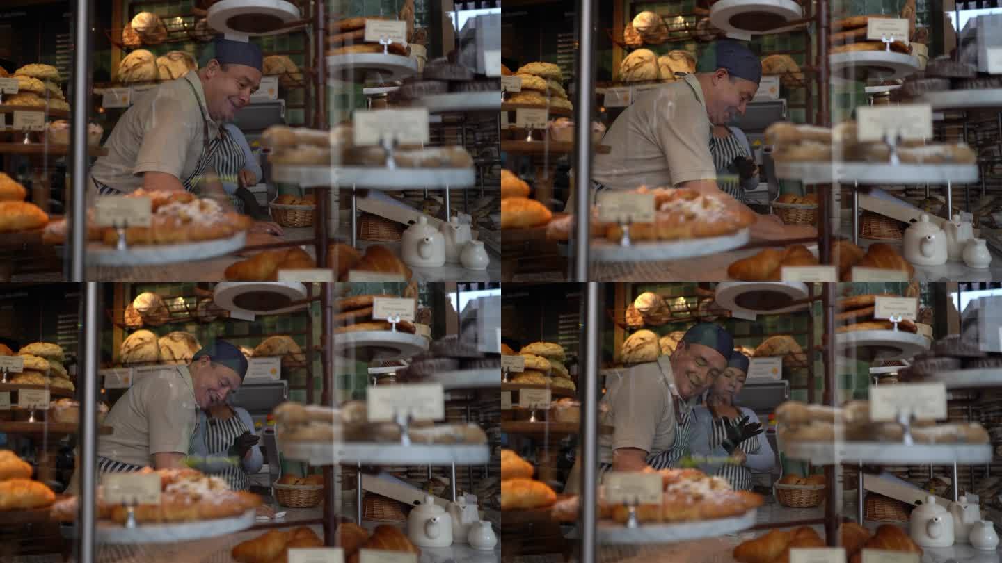 一个快乐的男人在一家面包店清洁柜台，而女人则戴着防护手套布置面包展示