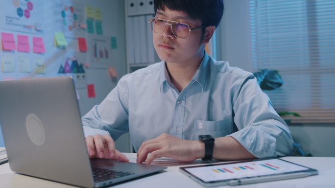 年轻的亚洲商人使用笔记本电脑查看屏幕滚动检查报告演示，并在小型工作场所晚上加班时比较数字平板电脑上的