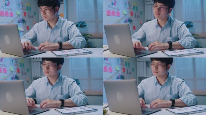 年轻的亚洲商人使用笔记本电脑查看屏幕滚动检查报告演示，并在小型工作场所晚上加班时比较数字平板电脑上的