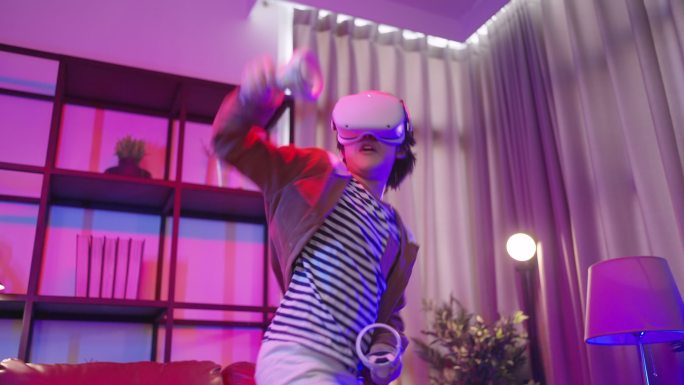 隔离期内，亚洲男童戴着VR或虚拟现实眼镜，戴着耳机站在家里的客厅里，在电视机前玩视频游戏，科技创新概