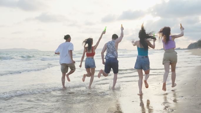一群亚洲年轻男女朋友一起在海滩上散步。迷人的旅行者在热带海岛度假旅行时，喝一瓶啤酒，感到快乐和放松