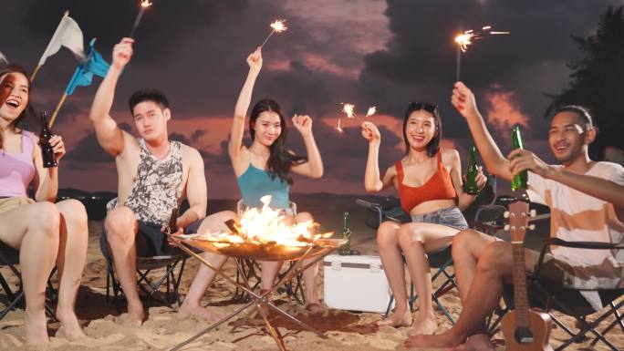一群亚洲年轻男女晚上在海滩上聚会。迷人的朋友，旅行者，一边在海边露营一边唱歌跳舞，一起在热带海岛享受