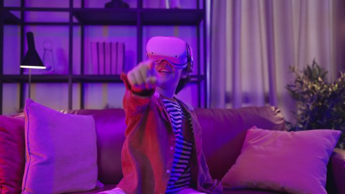 隔离期内，亚洲男童戴着VR或虚拟现实眼镜，戴着耳机站在家里的客厅里，在电视机前玩视频游戏，科技创新概