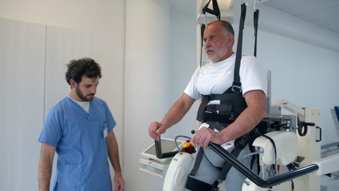 老年患者按照治疗师的指示，在物理康复中心，在电动外骨骼的帮助下，努力行走