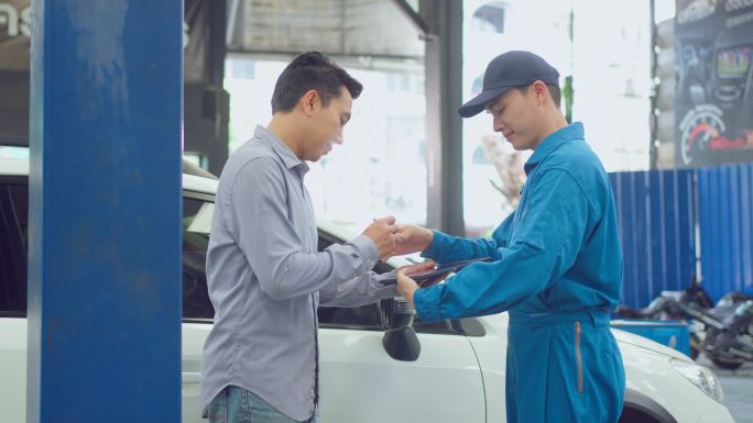 亚洲汽车技师在车库里向客户解释汽车状况。车辆服务经理男性在车辆部件的机械车间点工作，负责维护和修理汽