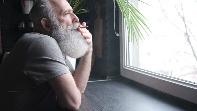 一位留着胡须的灰头发老人看着窗外，在厨房里抽烟