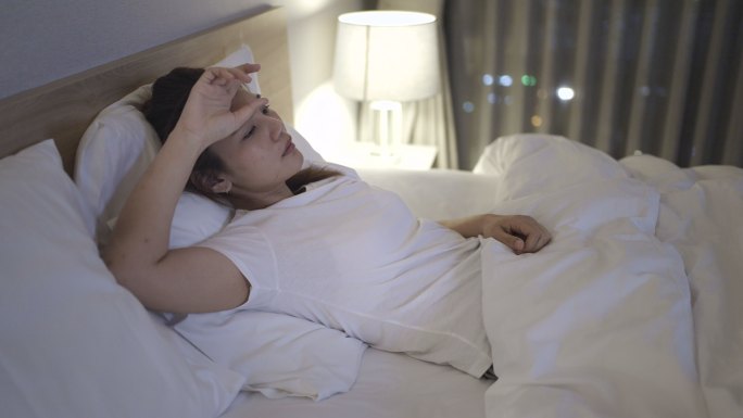 一位亚洲女性躺在卧室的白色床上，半夜在家里，看起来很焦虑，或者在思考自己的生活或工作。