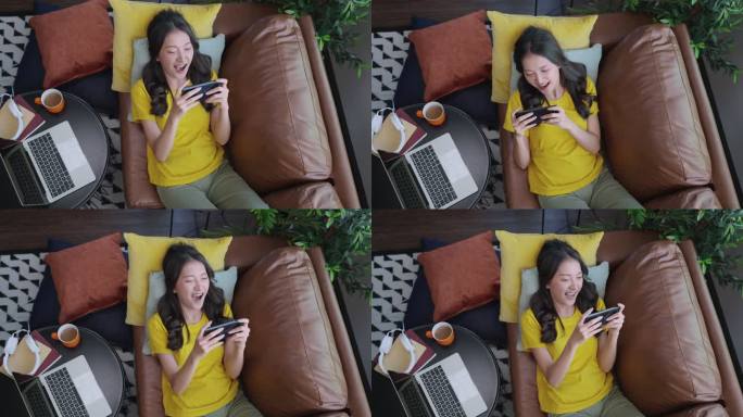 兴奋的微笑着的年轻成年亚洲女性戴着耳机，在家里沙发上用智能手机在线玩电子竞技游戏远程隔离封锁期家庭内