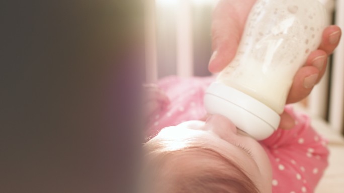 在阳光明媚的早晨，穿着粉色连体衣的可爱快乐女婴在室内喝婴儿奶瓶里的牛奶