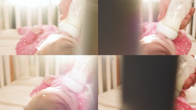 在阳光明媚的早晨，穿着粉色连体衣的可爱快乐女婴在室内喝婴儿奶瓶里的牛奶