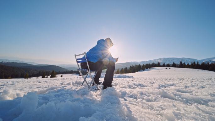 成熟的游客在网上冲浪，坐在雪地里的露营椅上。在大自然中独自在冬山露营。
