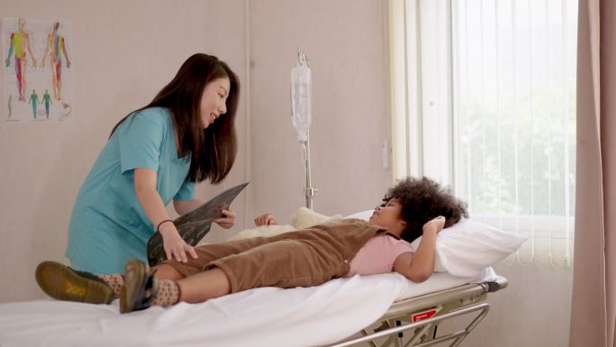 迷人的亚洲护士，拿着x光照片，与躺在病床上的儿童患者交谈，安慰他们，并用输液泵滴注生理盐水入院。