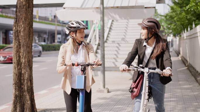 两位美丽的亚洲女商人戴着头盔，在人行道上散步，聊天。一个是骑自行车，另一个是拖着电动踏板车。可持续交