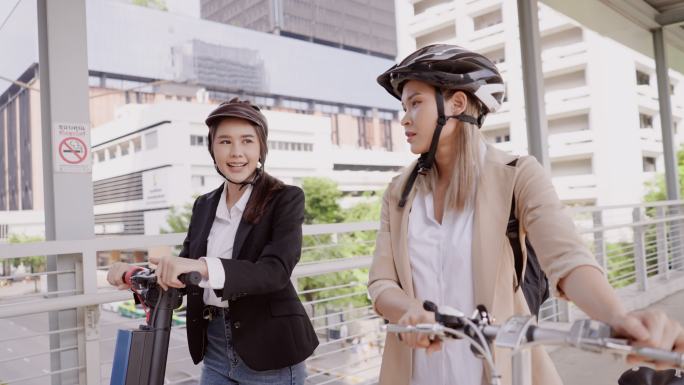 可持续流动性。两名年轻的亚洲女商人在穿过天桥前往办公楼时交谈。一位穿着棕色夹克的女商人正在拖着自行车
