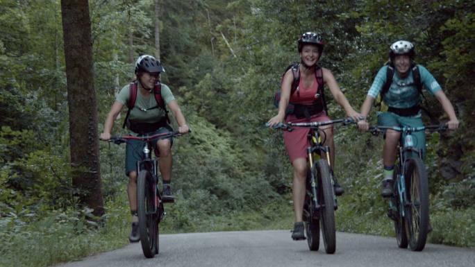 三名女子山地自行车手一起在树林中的一条铺好的道路上骑行
