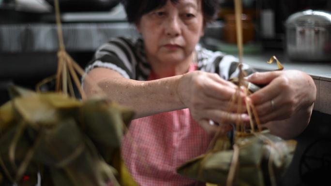 一位亚洲华裔高龄妇女在厨房里准备粽子，放入配料并将其包装起来，为端午节做准备