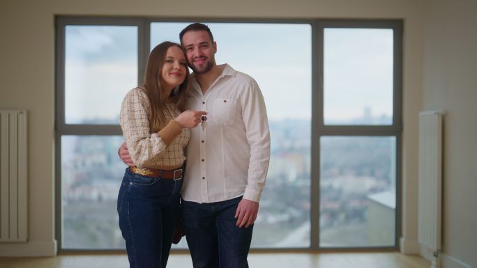 年轻夫妇拿着新家的钥匙，在新家里对着相机微笑的照片