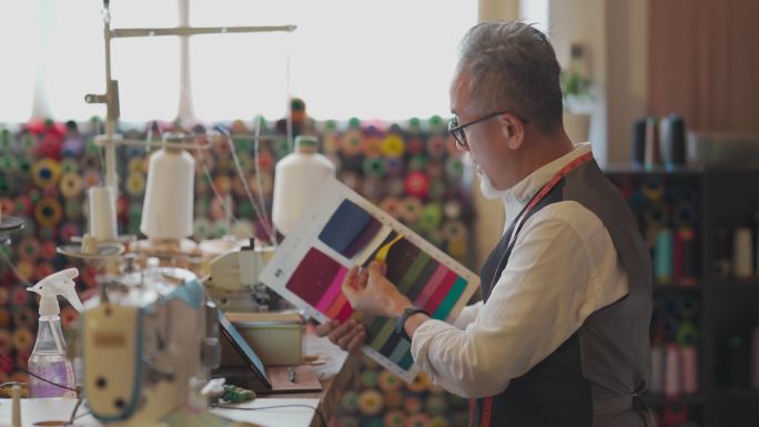 一名亚裔中国高级男子在工作室展示变种纺织面料样本目录视频，用数字平板电脑与客户通话