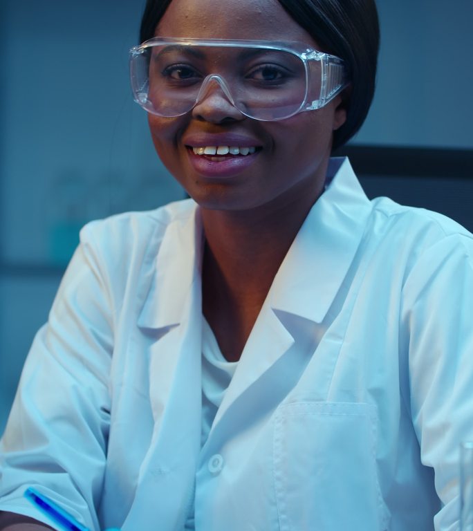 一位美丽的非裔美国医生坐在现代化实验室的工作场所，面带微笑地看着摄像机