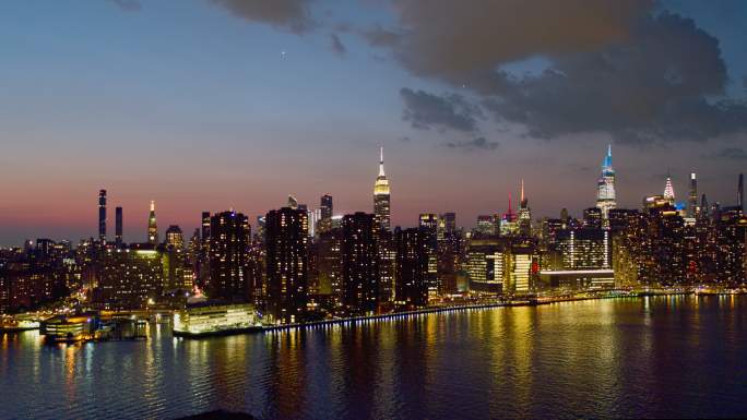 日落时分，纽约市曼哈顿中城东河对岸的远处全景夜景。向后移动摄像机的空中视频。