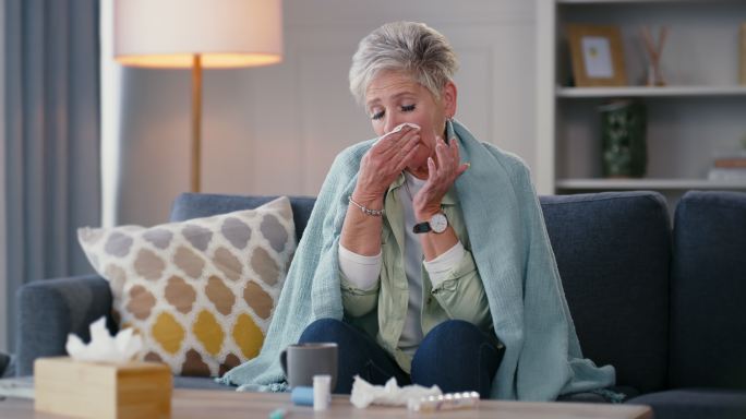 生病的老太太用纸巾擤鼻涕。在家客厅里，一位患有鼻窦、感冒、流感和过敏症的成熟退休女士。试着保暖，盖上