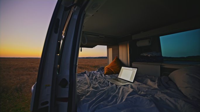 黄昏时分，停在乡下某处的露营车驾驶室内的床上的DL笔记本电脑