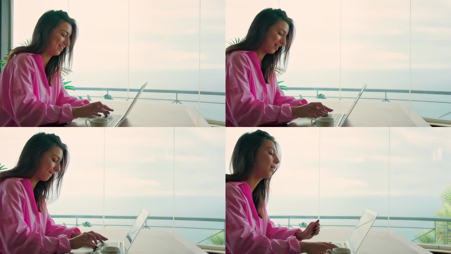 在全景窗边工作的快乐女人。使用笔记本电脑，喝咖啡欣赏壮丽的海景