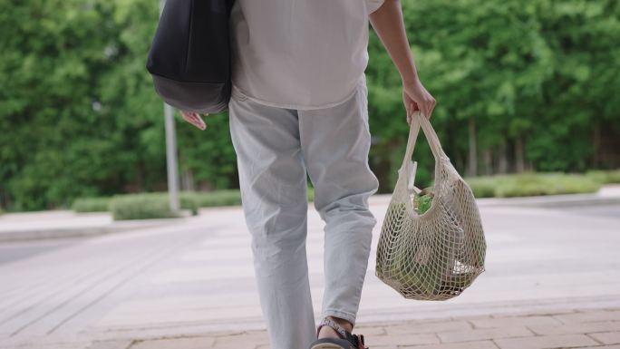 穿着休闲服的年轻女子手持可重复使用的网袋穿过绿色城市场景中的道路