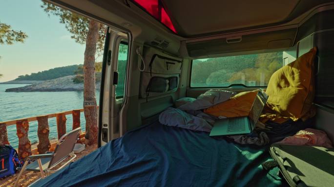 在一辆货车里面，笔记本电脑放在床上，枕头和毯子，货车停在海边，外面的露营椅，货车生活