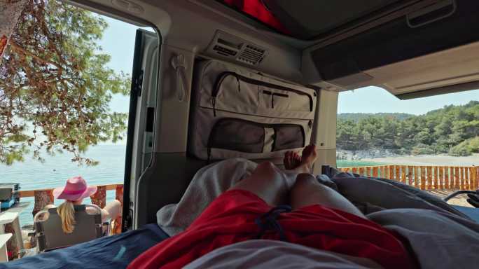 一名男子躺在开着门的面包车里，他的金发妻子戴着粉色帽子坐在外面的露营椅上晒太阳