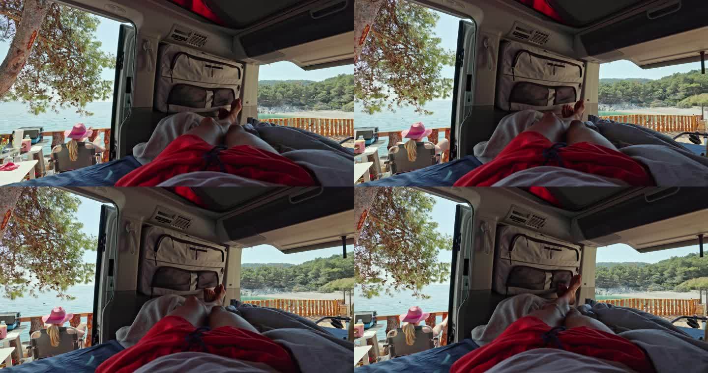一名男子躺在开着门的面包车里，他的金发妻子戴着粉色帽子坐在外面的露营椅上晒太阳