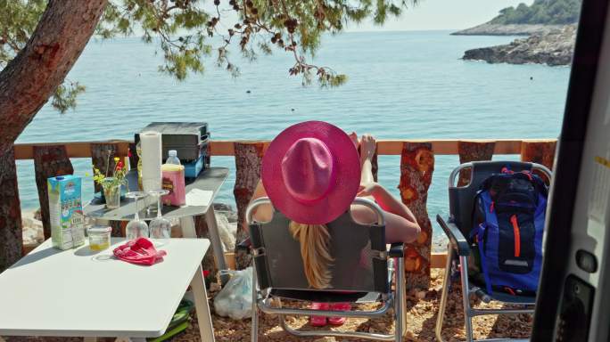 戴着粉红色帽子的金发女人在阳光下放松，坐在一张露营椅上，双腿伸开，面包车内的后视图，《面包车生活》