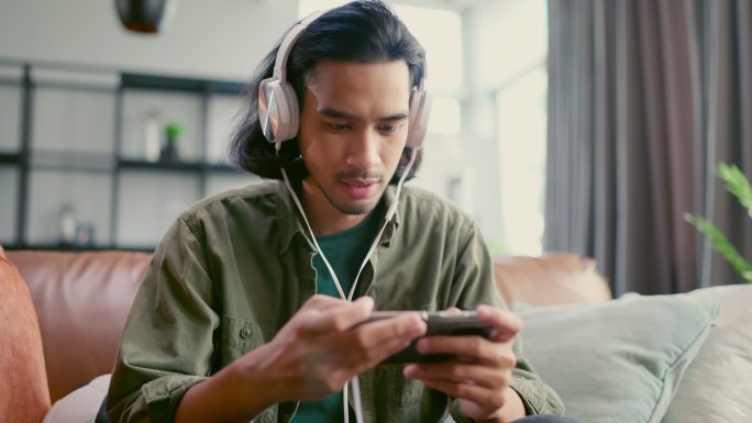 亚洲男性玩手机游戏。上瘾男人。虚拟娱乐。休闲男子坐在沙发沙发客厅里，在智能手机上玩网络游戏。