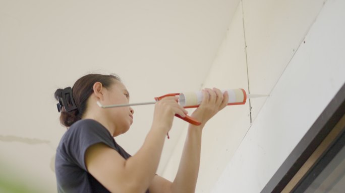 一名亚洲女性爬上梯子，用硅胶枪封住腐烂的墙壁接缝，以掩盖裂缝。女性工作人员在家里腾出新房间或在家里翻