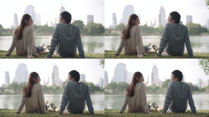 一对亚洲情侣坐在绿草地上聊天，与美丽的城市景观人们在城市自然环境中放松。