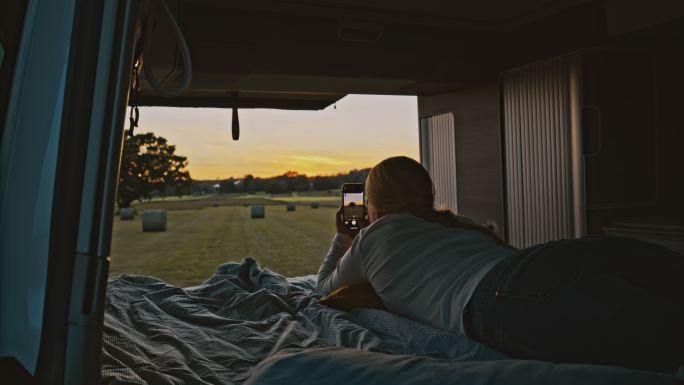 DL Woman在日落时分停在草地上的一辆露营车的小屋里用手机拍照