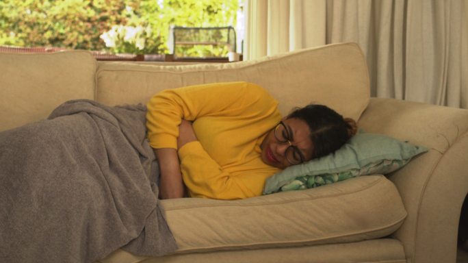 一位年轻女子躺在家里的沙发上，胃痛。胃痛的女人，拿着毯子躺在沙发上