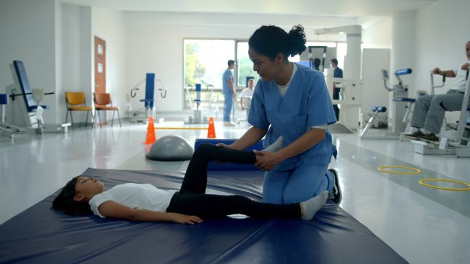 小女孩躺在体操垫上，在物理康复中心进行理疗，帮助她伸展双腿