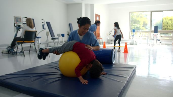 男孩在理疗师的帮助下，在健身球上进行体育锻炼